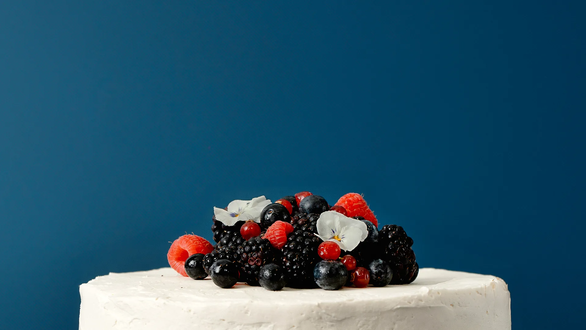 Bolo decorado com frutas vermelhas frescas e flores comestíveis sobre fundo azul. Delicioso e elegante.