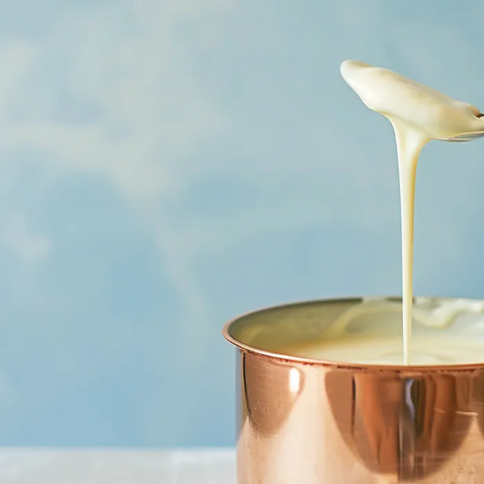 Creme branco escorrendo de uma colher em panela de cobre, fundo azul claro.