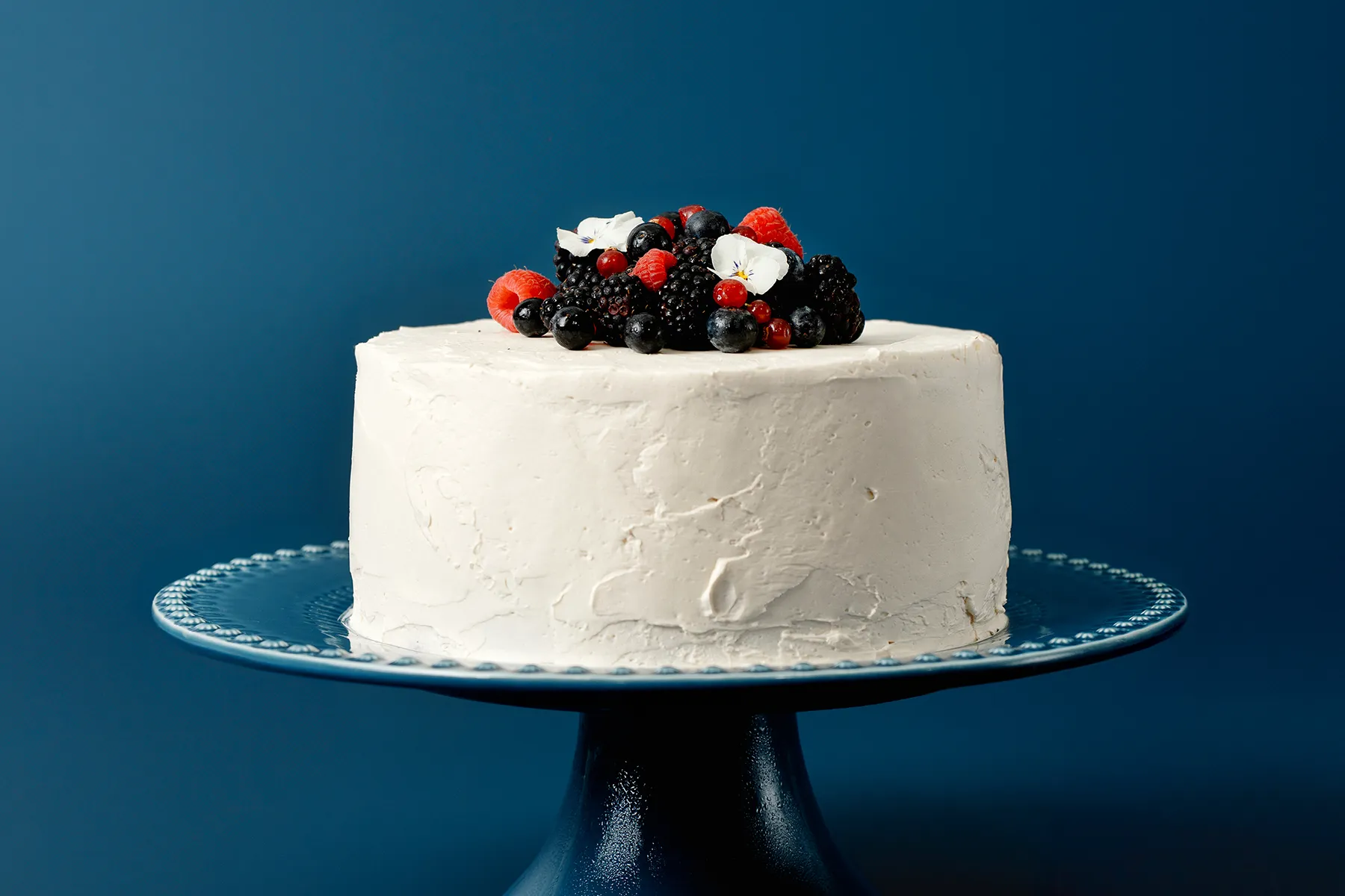 Bolo branco decorado com frutas vermelhas e flores comestíveis em prato azul com fundo azul.