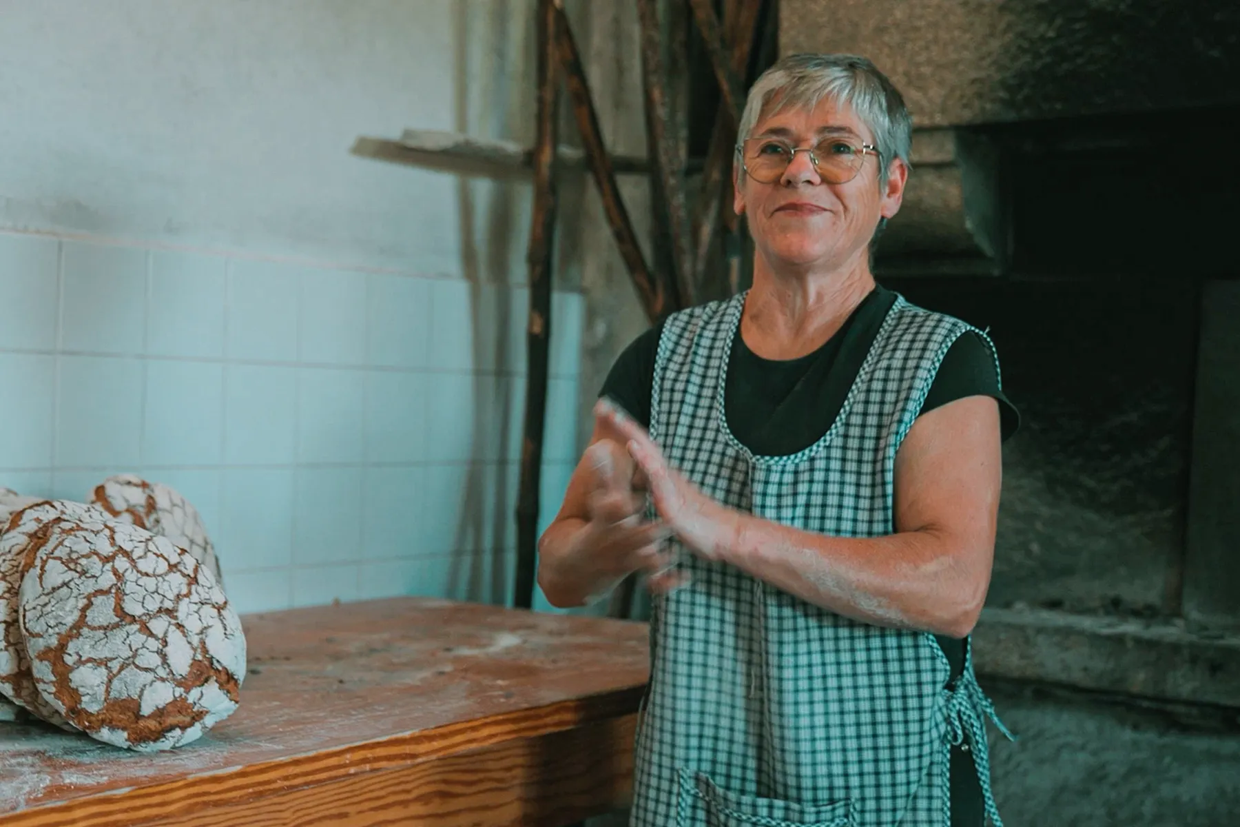 Padeira sorrindo em frente a forno tradicional de pães rústicos. Ambiente acolhedor de padaria artesanal.