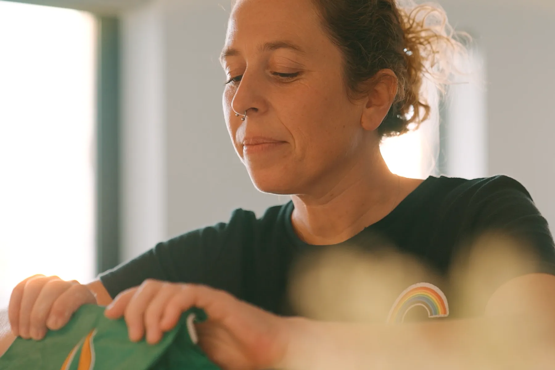 Mulher concentrada abrindo embalagem verde, com luz suave ao fundo e detalhe de arco-íris na camisa.