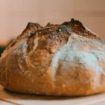 Pão artesanal recém-assado sobre tábua de madeira na cozinha, com crosta dourada e textura rústica.